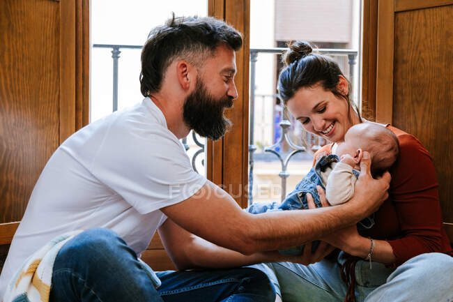 Barbuto padre e madre felice comunicare con bambino carino mentre seduto sul pavimento vicino alla finestra a casa — Foto stock