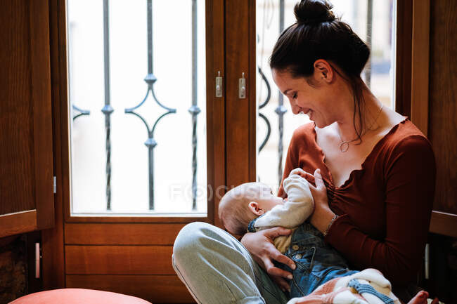 Cheerful woman hugging and breastfeeding cute baby while sitting near window and resting in cozy room at home — Stock Photo