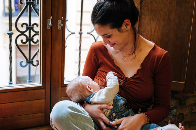 Happy mother breastfeeding baby near window — Stock Photo