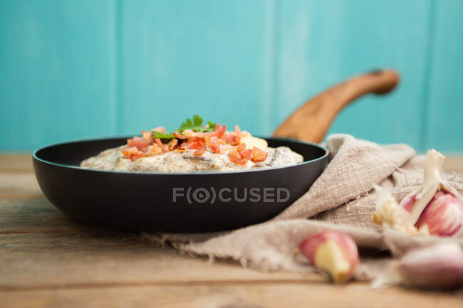 Composition of tasty spaghetti with ham slices and mushrooms in creamy sauce cooked in pan and placed on wooden cutting board at a wooden table with garlic and linen fabric aside on blue background — Stock Photo