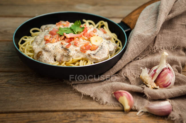 De haut savoureux spaghettis avec tranches de jambon et champignons en sauce crémeuse cuite dans une casserole et placée sur une planche à découper en bois à une table en bois avec de l'ail et du tissu de lin de côté — Photo de stock
