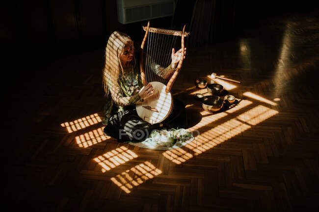 Зверху доросла жінка сидить на сонячному світлі біля співочих мисок і грає традиційну мелодію на лірі в темній кімнаті вдома — стокове фото