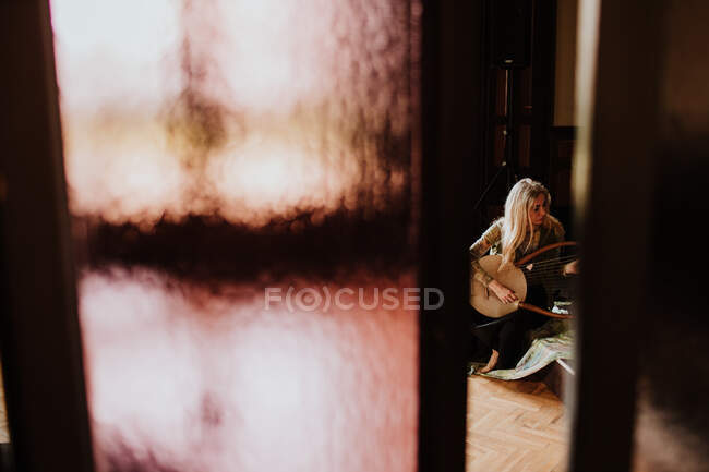 Mulher adulta tocando melodia tradicional na lira enquanto sentada no chão atrás da partição no quarto acolhedor em casa — Fotografia de Stock