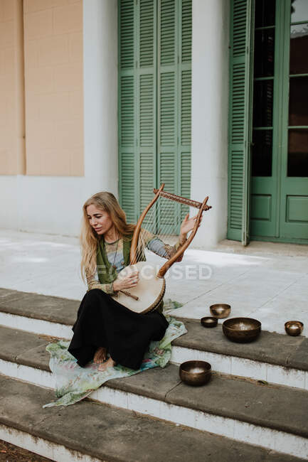Full body woman sitting on building stairs near tibetan bowls and playing lyre on street — Stock Photo