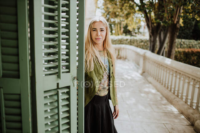 Adult woman with blond hair looking at camera while standing near old building entrance on marble terrace on sunny day in garden — Stock Photo