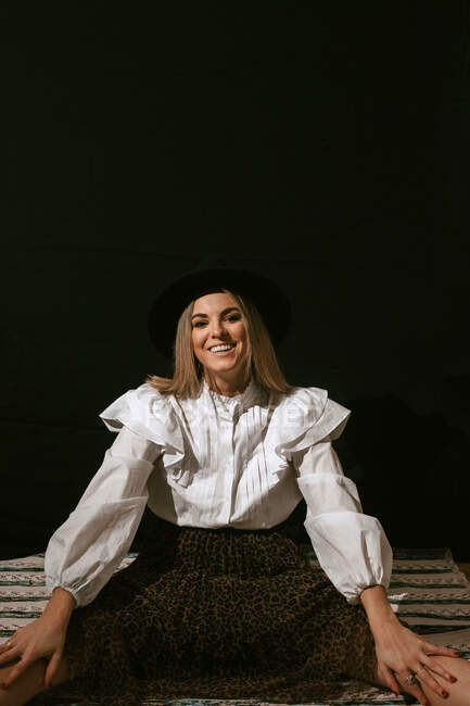 Cheerful young cute blonde female in stylish outfit and hat sitting on floor and smiling for camera against black background — Stock Photo