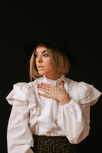 Jovem mulher loira bonito em roupa elegante e chapéu olhando para longe contra o fundo preto — Fotografia de Stock