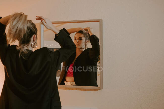 Jovem mulher em roupas elegantes olhando para o espelho e fazendo rabo de cavalo de cabelo loiro em casa — Fotografia de Stock
