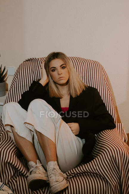 Menina na moda com cabelo loiro sentado em poltrona listrada e olhando para a câmera enquanto descansa em casa — Fotografia de Stock