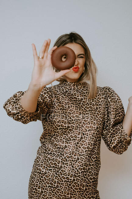 Jovem fêmea de vestido com leopardo imprimir lábios bebendo e olhando para a câmera através de doce donut de chocolate contra fundo cinza — Fotografia de Stock