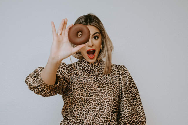 Jeune femme blonde mignonne en robe avec imprimé léopard boudant les lèvres et regardant la caméra à travers un beignet au chocolat doux sur fond gris — Photo de stock