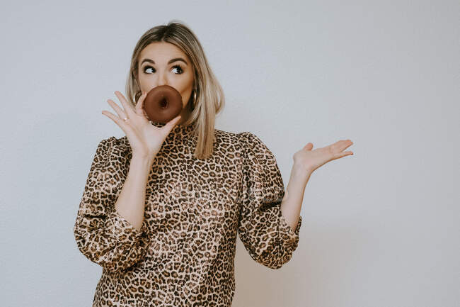 Young blonde female in a leopard print dress pouting and covering lips with chocolate doughnut looking away while standing against gray background — Stock Photo