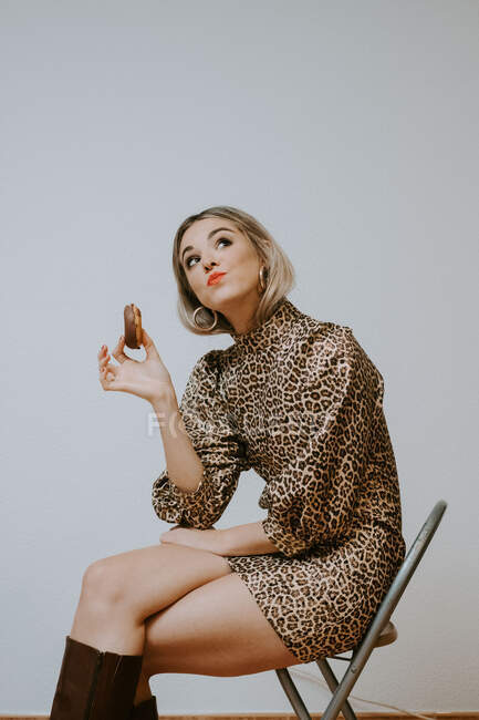 Jovem mulher loira feliz em vestido da moda com leopardo impressão sorrindo olhando para longe mordendo saboroso donut de chocolate enquanto sentado na cadeira contra fundo de parede cinza — Fotografia de Stock