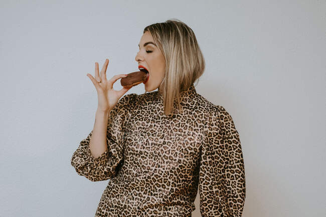 Glückliche junge blonde Frau im trendigen Kleid mit Leopardenmuster lächelt mit geschlossenen Augen und beißt leckeren SchokoladenDonut, während sie vor grauem Hintergrund steht — Stockfoto