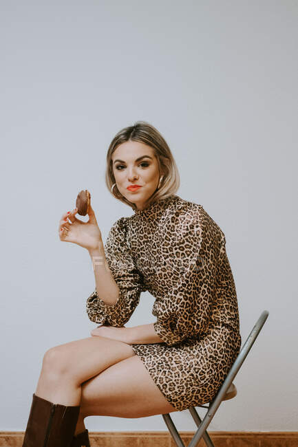 Щаслива молода блондинка в модній сукні з леопардовим принтом, усміхнений, дивлячись на камеру, кусає смачний шоколадний пончик, сидячи на стільці на сірому фоні стіни — стокове фото