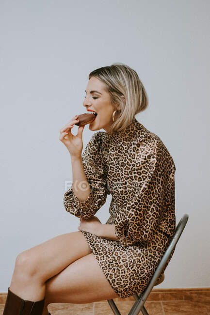 Feliz joven rubia en vestido de moda con estampado de leopardo sonriendo con los ojos cerrados mordiendo sabrosa rosquilla de chocolate mientras está sentado en la silla contra fondo de pared gris - foto de stock