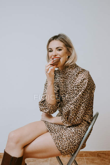 Felice giovane donna bionda in abito alla moda con stampa leopardo sorridente guardando la fotocamera mordere gustosa ciambella al cioccolato mentre seduto sulla sedia contro sfondo muro grigio — Foto stock