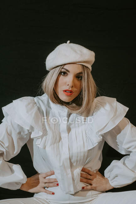 Attraktive junge Frau in trendiger weißer Kleidung und Baskenmütze hält die Hände auf der Taille und blickt vor schwarzem Hintergrund in die Kamera — Stockfoto
