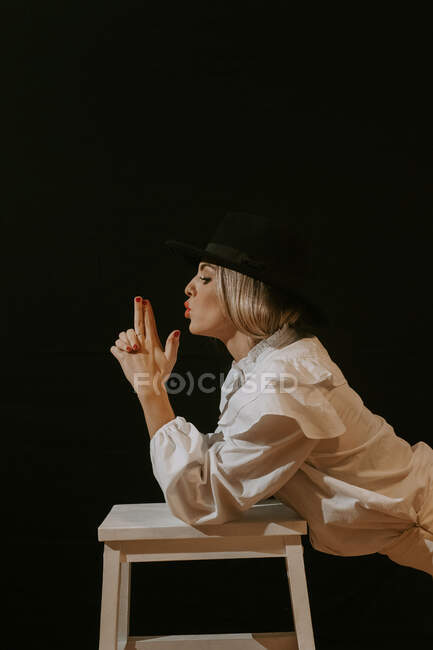 Side view of young blonde woman in white blouse and stylish hat looking away while leaning on stool blowing and making gun gesture with fingers and against black background — Stock Photo