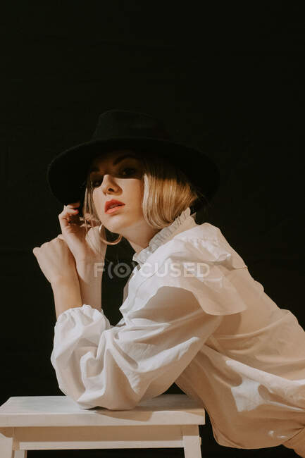 Seitenansicht einer jungen blonden Frau in weißer Bluse, die in die Kamera schaut und ihren eleganten Hut anpasst, während sie sich auf einen Hocker vor schwarzem Hintergrund lehnt — Stockfoto