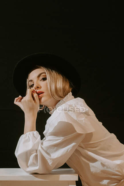Seitenansicht einer jungen blonden Frau in weißer Bluse und stylischem Hut, die in die Kamera blickt, während sie sich auf einen Hocker vor schwarzem Hintergrund lehnt — Stockfoto