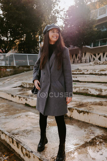 Щаслива молода жінка в модному верхньому одязі посміхається і дивиться геть, стоячи на посушливих сходах в сонячний осінній день в парку — стокове фото