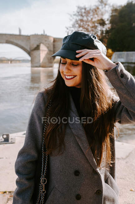 Happy young woman in trendy outerwear smiling with closed eyes and adjusting cap while standing on quay near river on sunny day — Stock Photo