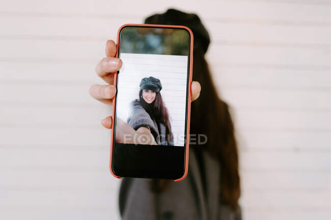 Verschwommene junge Frau demonstriert Smartphone mit Selfie, während sie an Hauswand auf der Stadtstraße steht — Stockfoto