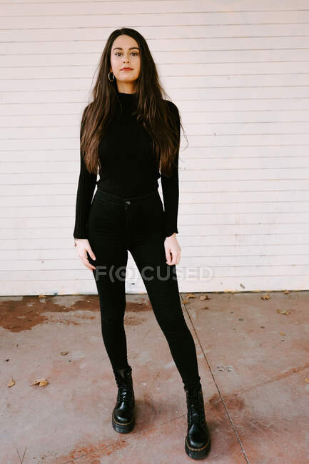 Все тело молодая женщина в модном черном наряде стоит перед белым зданием и смотрит в камеру на городской улице — стоковое фото