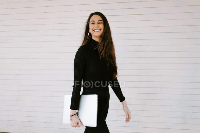 Jovem alegre com laptop moderno sorrindo e olhando para longe enquanto caminhava contra a parede branca na rua da cidade — Fotografia de Stock