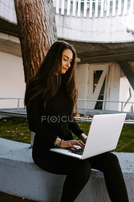 Young woman in trendy black outfit typing on laptop keyboard while sitting on border near tree in courtyard of modern building — Stock Photo