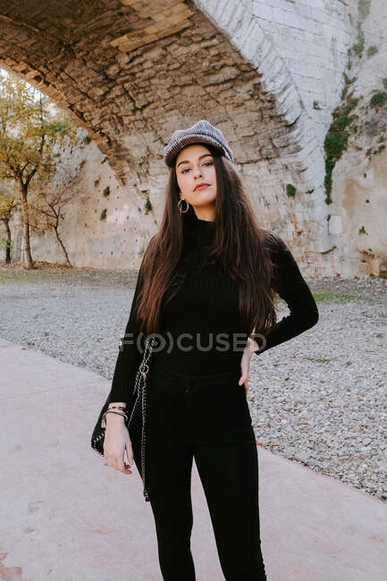 Young woman in stylish outfit and cap keeping hand on waist and looking at camera while standing against old arch in park — Stock Photo
