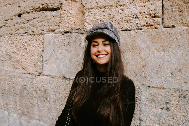 Happy young woman in trendy hat smiling and looking at camera while standing near shabby wall of aged stone building — Stock Photo