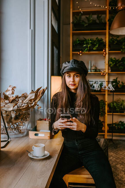 Giovane donna in abiti eleganti guardando la fotocamera e la navigazione smartphone mentre seduto vicino alla finestra in accogliente ristorante — Foto stock