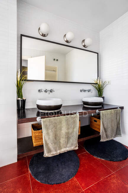 Banheiro moderno com lâmpadas e grande espelho colocado sobre balcão elegante com pias e tapetes redondos acolhedores no piso vermelho — Fotografia de Stock