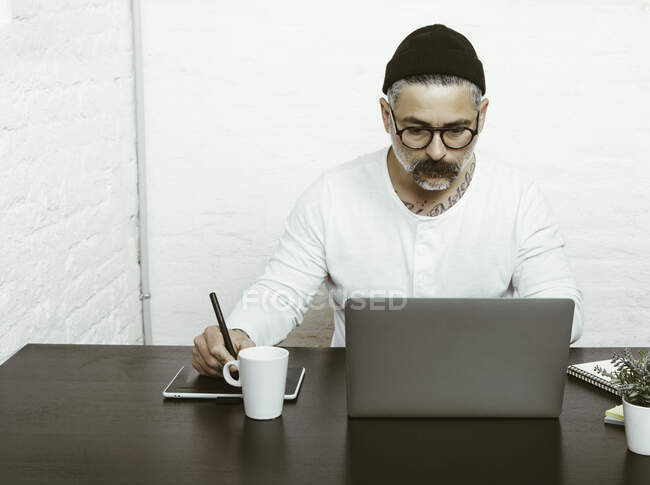 Концентрований бородатий чоловік в окулярах і головних уборах використовує нетбук для роботи вдома в карантинний час — стокове фото