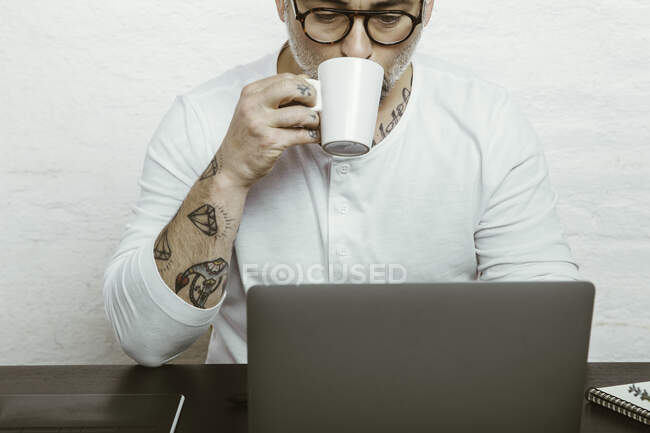 Focused male in glasses with tattoos on arms drinking coffee and surfing laptop while working at home because of quarantine — Stock Photo