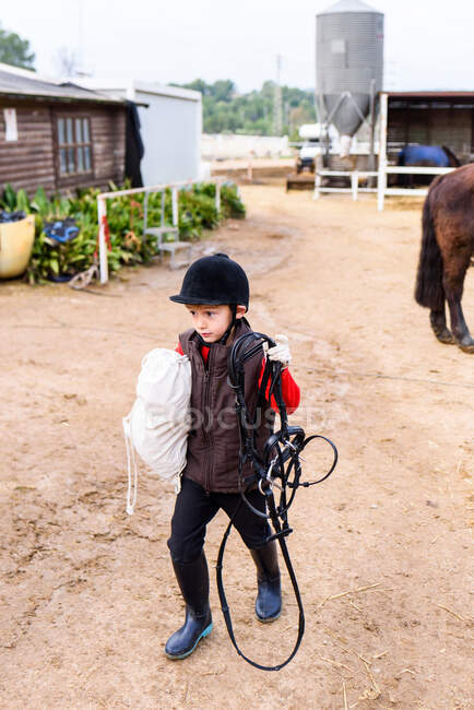 Little jockey carrying equipment on arena after training in equestrian school — Stock Photo