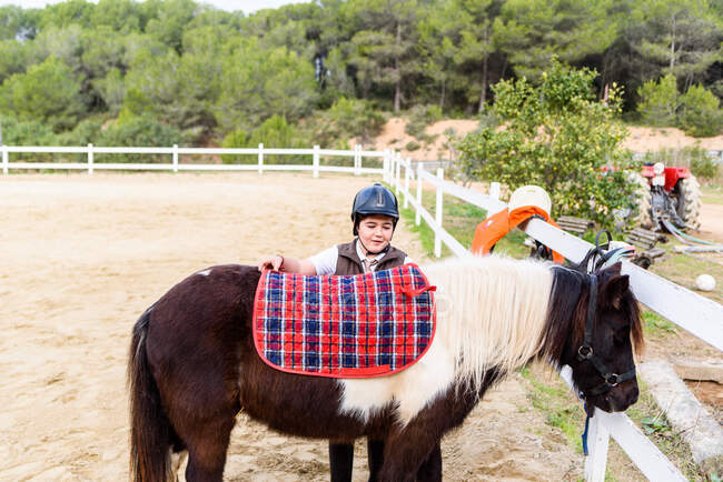 Little jockey in protective helmet adjusting stirrup on saddle before riding skewbald pony in equestrian school — Stock Photo
