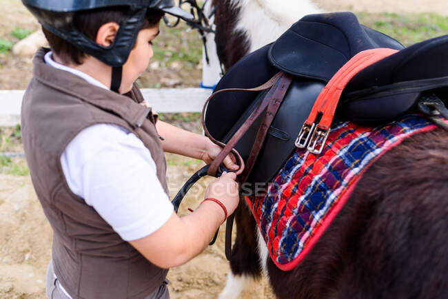 Vista lateral de pouco jockey no capacete de proteção ajustando estribo na sela antes de montar pônei skewbald na escola equestre — Fotografia de Stock
