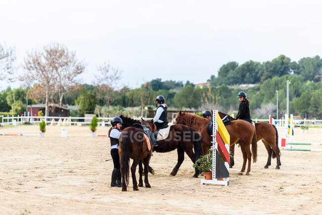 Teen jockeys in helmets communicating with each other while riding obedient horses on sandy dressage arena during lesson in equestrian school — Stock Photo