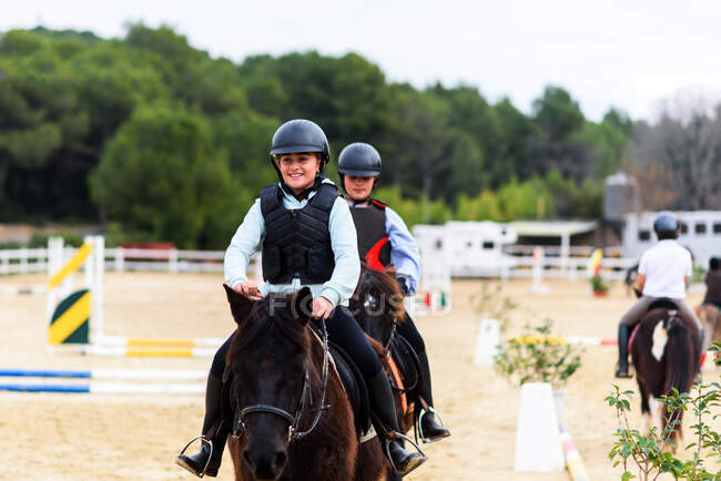 Happy teen jockeys in helmets riding obedient horses on sandy dressage arena during lesson in equestrian school — Stock Photo