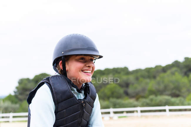 Chica adolescente alegre en casco de jinete y chaleco sonriendo y mirando hacia otro lado durante la clase de equitación en la escuela ecuestre en el campo - foto de stock