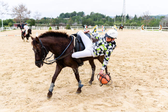 Adolescente jockey jogar com bola em solo arenoso de volta de cavalo marrom durante a lição na escola equestre — Fotografia de Stock