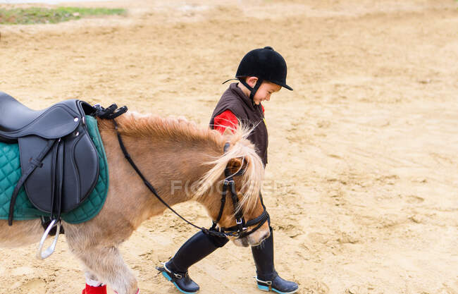 Вид збоку веселого хлопчика в костюмі хокею і шоломі, що веде поні рани під час прогулянки на піщаному ґрунті виїзної арени в кінній школі — стокове фото