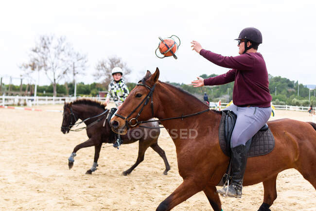 Adolescentes chicos en cascos lanzando pelota entre sí mientras montan caballos en la arena de doma durante la lección en la escuela ecuestre - foto de stock