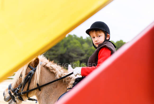Vista lateral del jinete pequeño en casco mirando a la cámara mientras monta pony con melena trenzada detrás de la barrera roja y amarilla en la arena de doma - foto de stock