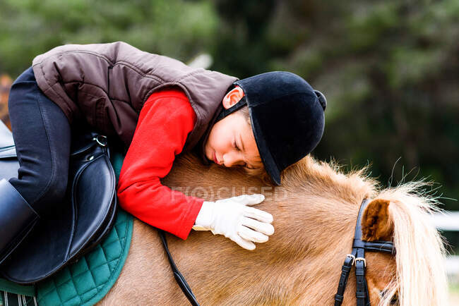 Vista lateral da criança com olhos fechados no traje de jóquei sentado em sela e abraçando pônei roan durante a aula na escola de equitação — Fotografia de Stock