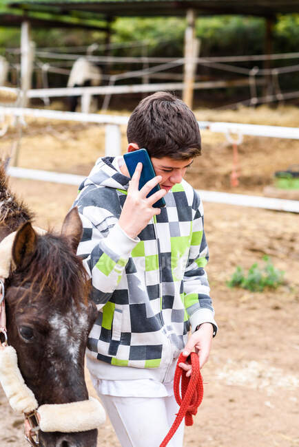 Menino adolescente com capuz quadriculado levando cavalo obediente e tendo uma conversa de smartphone enquanto caminha no paddock durante o treinamento na escola equestre — Fotografia de Stock