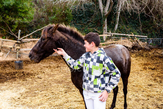 Allegro ragazzo adolescente in felpa con cappuccio a scacchi sorridente e toccante testa di cavallo marrone mentre in piedi nel paddock dopo l'allenamento nella scuola equestre — Foto stock